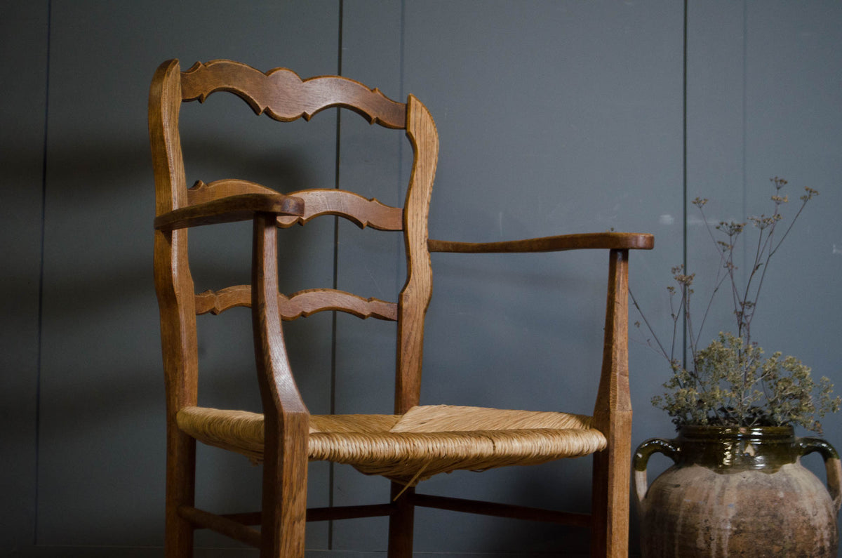 A wooden armchair with a woven rope seat and a curved backrest, positioned next to a rustic ceramic vase with dried stems against a dark gray paneled wall.