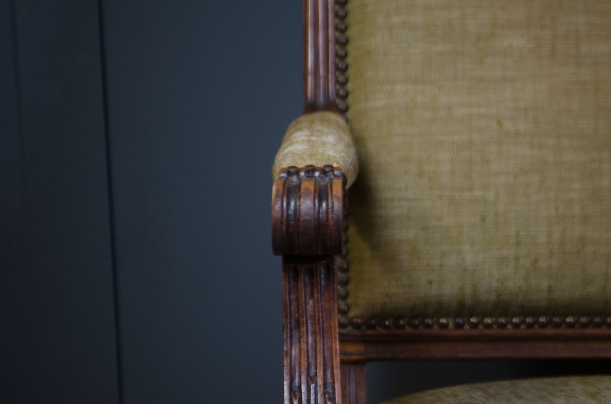 
Close-up detail of an antique wooden armchair, showing the carved dark brown wood frame, decorative nailhead trim, and textured beige fabric upholstery.