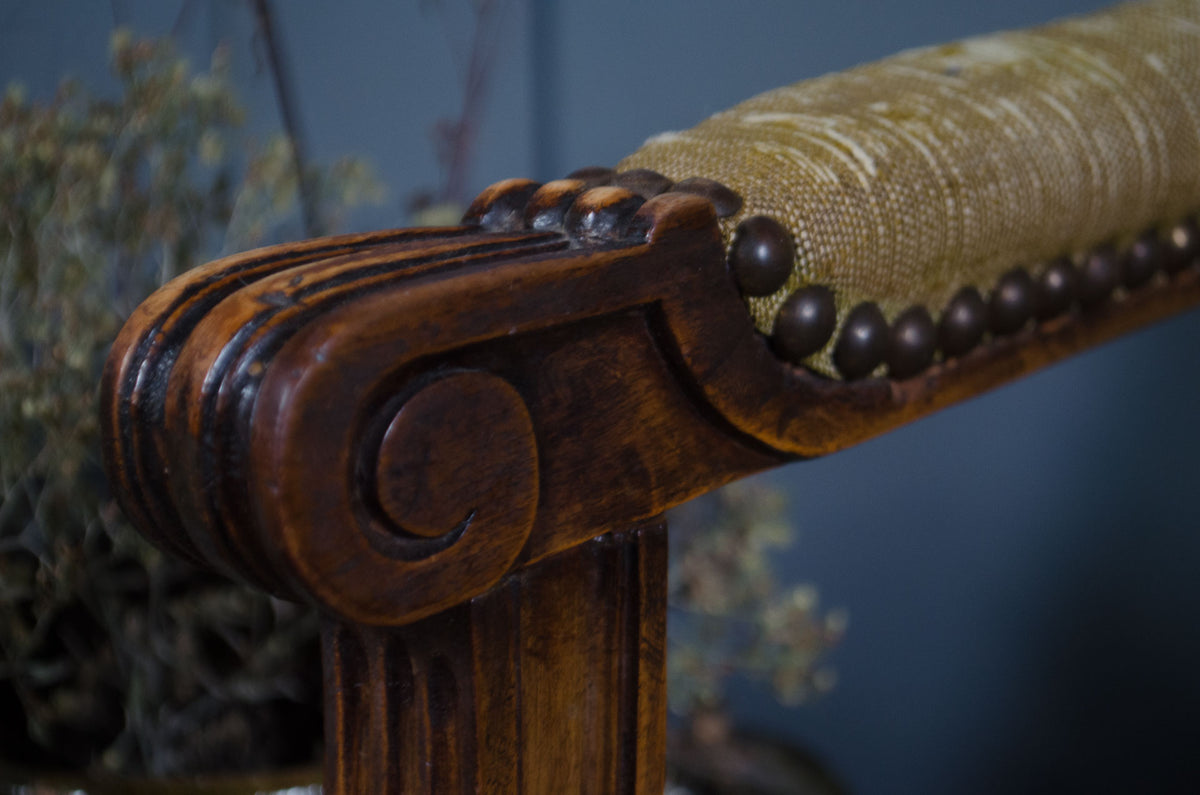 Close-up of an antique wooden chair arm with ornate carved wood detailing, dark nailhead trim, and a textured beige fabric upholstery, set against a dark blue background.