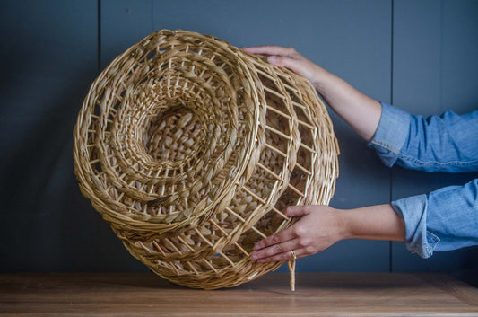 A person's hands holding a large, round, woven wicker basket with a spiral pattern, set against a dark blue paneled wall and a wooden table.