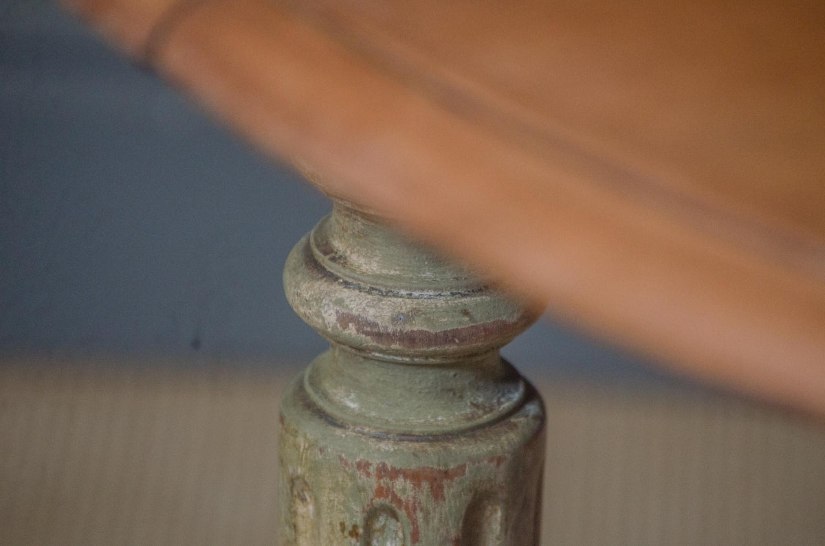 
Close-up of a distressed painted wooden table leg with a weathered green and red finish, partially obscured by a warm brown wooden tabletop in the foreground.