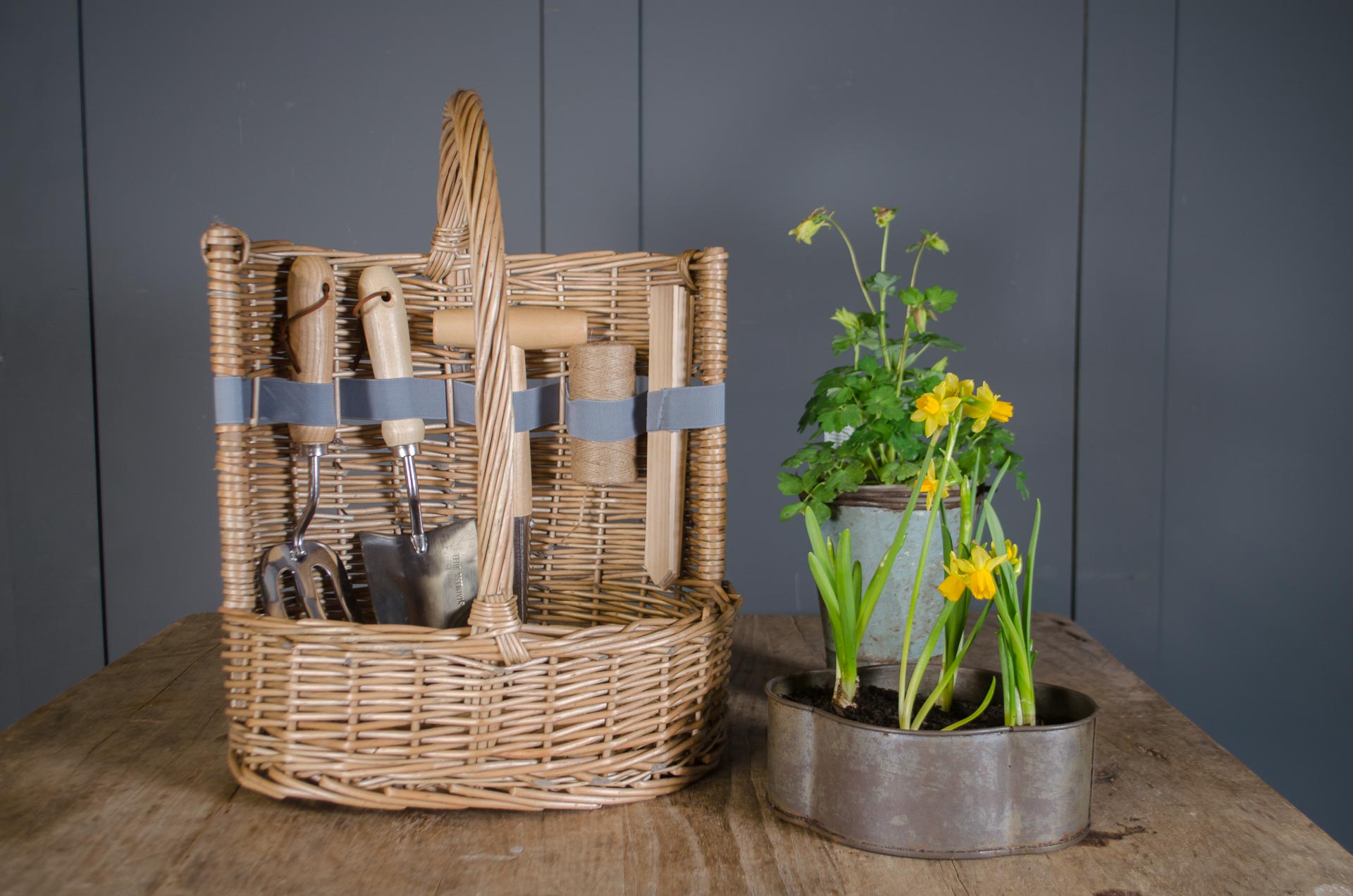 
Wicker garden trug basket with wooden handled tools including trowel, fork, dibber and twine displayed on table