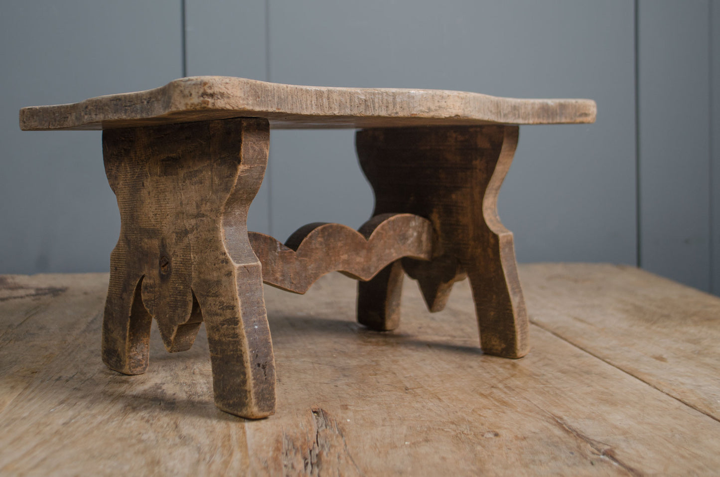 Primitive French wooden stool with weathered grey finish and carved rustic base on wooden table