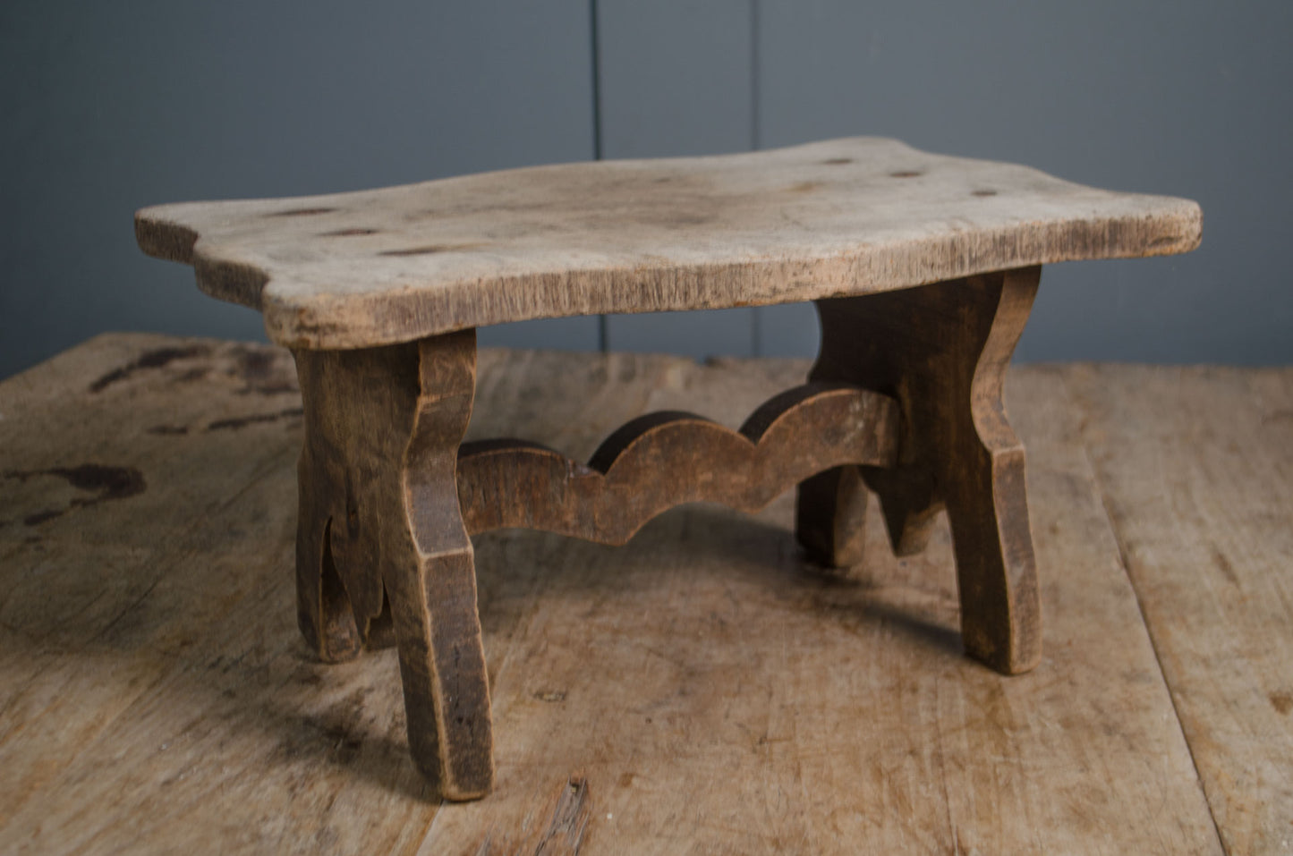 Primitive French wooden stool with weathered grey finish and carved rustic base on wooden table