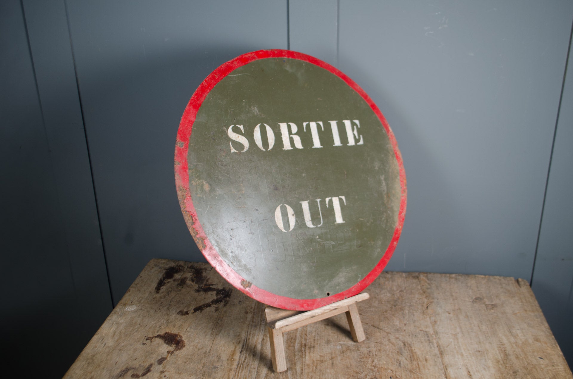 Round sign with 'SORTIE OUT' text on a stand against a gray wall.