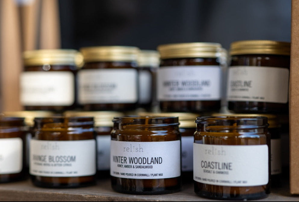 A collection of amber glass jars with gold aluminium lids, containing hand-poured candles in various fragrances. The jars are labelled with their fragrance names.